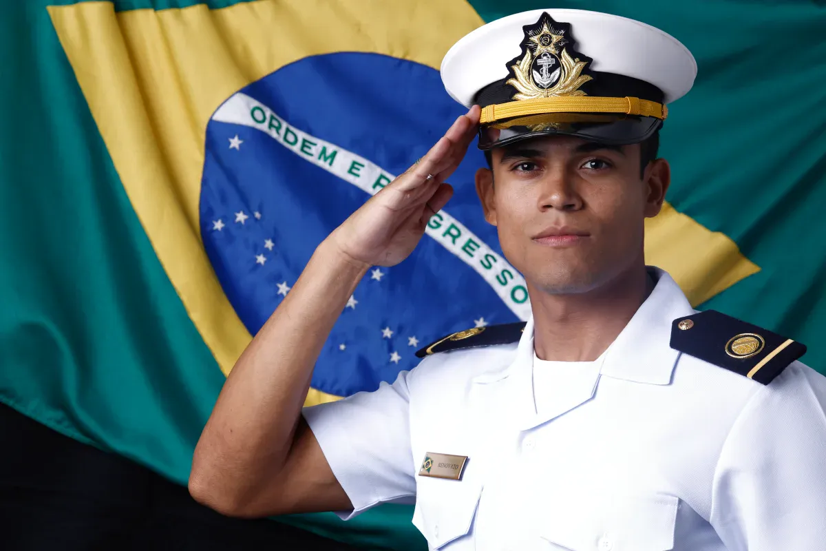 Matheus Renovato de farda branca da Marinha, batendo continência, com a bandeira do Brasil ao fundo